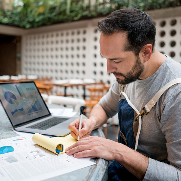 A man writes notes down while an open laptop shows various charts.