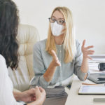 Masked woman talks to another woman in an office.