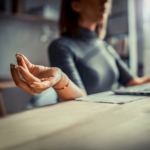 A close-up shot of a woman touching each index finger to each thumb in a yoga “mudra” gesture while sitting in front of a laptop.