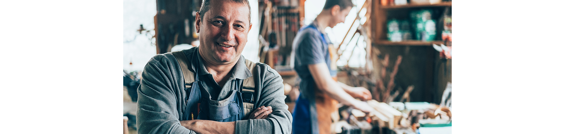 The owner of a small business smiles at a camera. In the background is his son, who is busy at work.