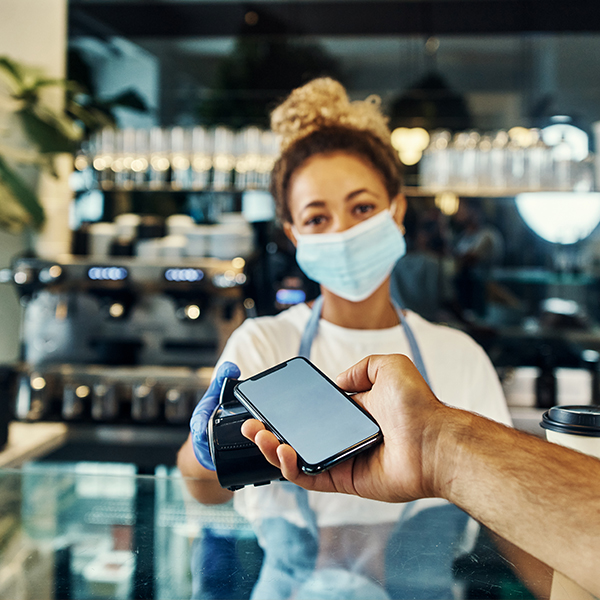 A masked woman behind a counter receives contactless payment from a smartphone.