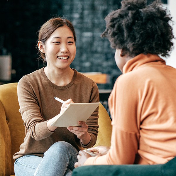 Two women, one taking notes, have a discussion in an office lounge.