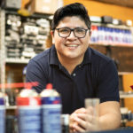 A small business owner smiles behind the counter of a hardware store while an employee stocks shelves in the background.