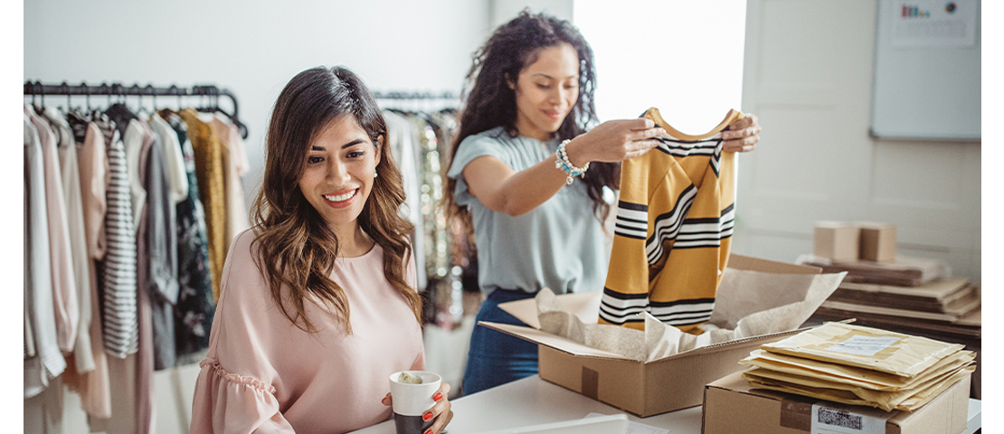Two women in the clothing store, one is packaging a clothes in the box and the other is holding a cup of drink