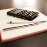 A calculator, a pencil, and a pen lie on a blank notepad.