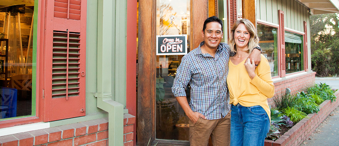 A man and woman smile next to each other in front of their small business.