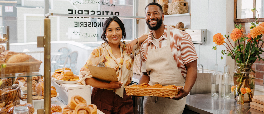 A man holds a basket of bread while a woman next to him cradles a clipboard in a bakery.